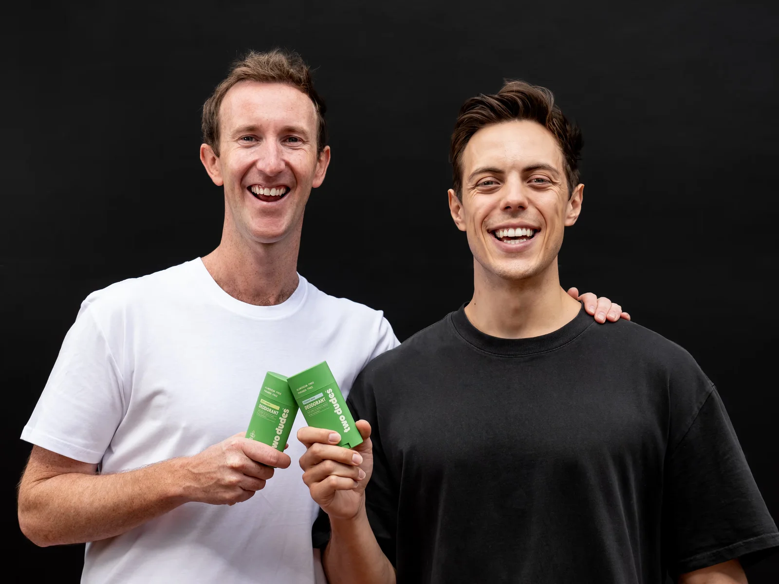 Two Dudes founders holding the green deodorant sticks against a black studio backdrop